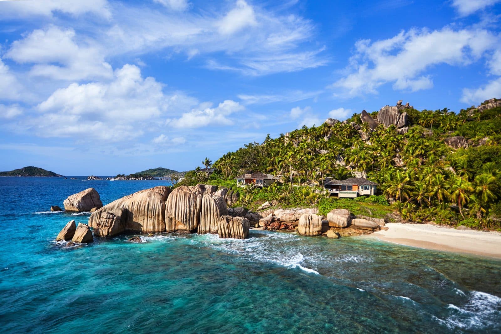 Granite boulders at the edge of turquoise water, a beach and villas among palms on Félicité Island's hillside