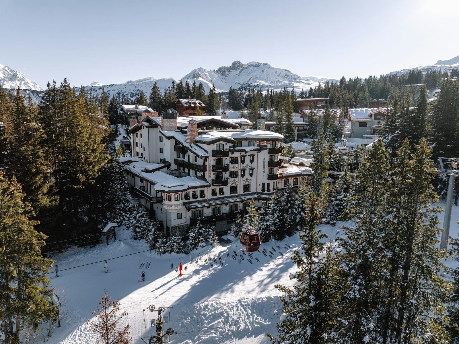 Aerial view of Les Airelles: a white turreted building with a painted façade among fir trees, a piste and lift cabin below, and mountains on the horizon