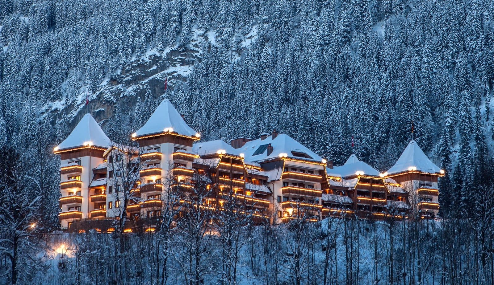 The Alpina Gstaad on a winter evening: a turreted building with snowy roofs and glowing balconies among a snowy fir forest