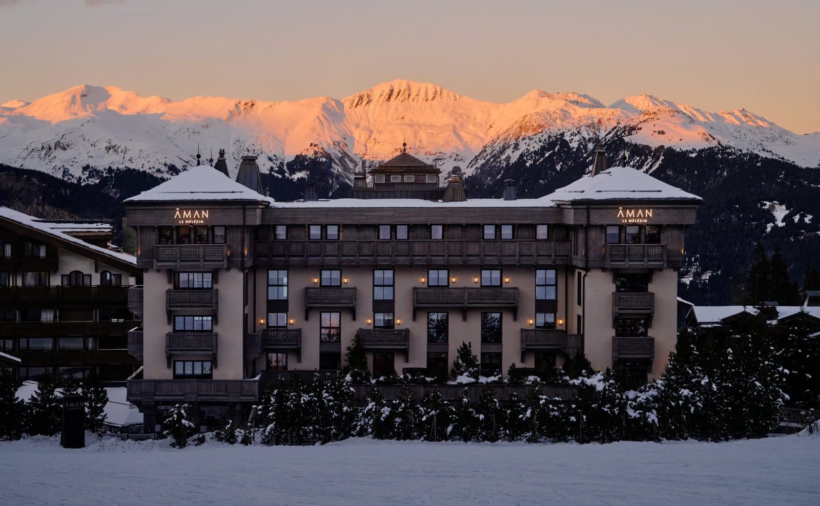 The façade of Aman Le Mélézin at dusk: glowing windows, snowy fir trees below and peaks in pink sunset light behind the hotel