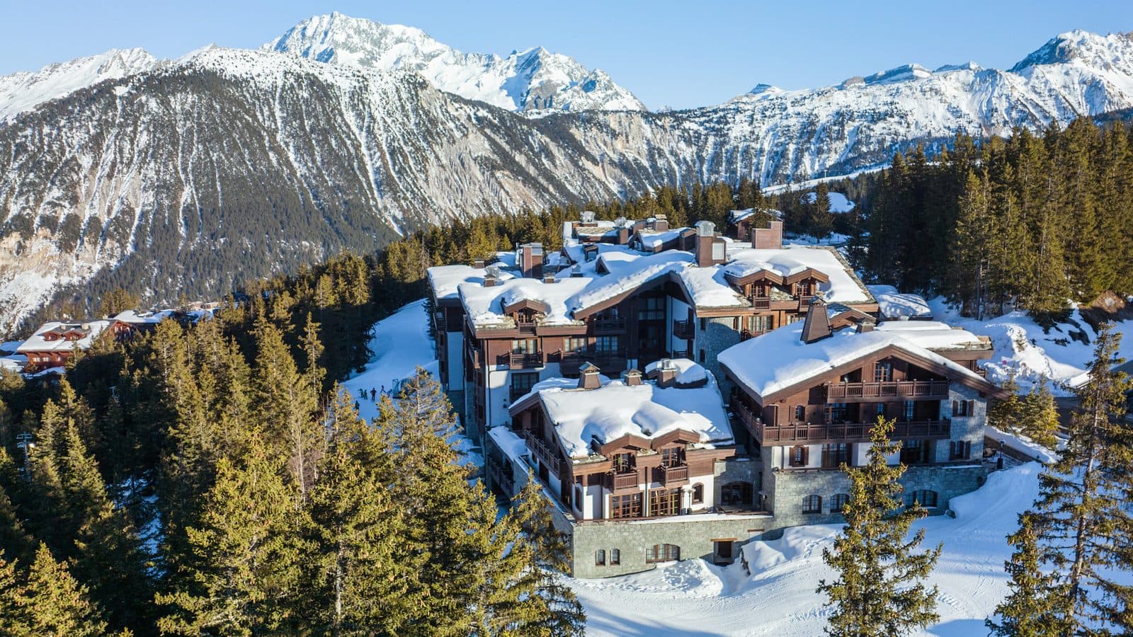 Aerial view of L’Apogée Courchevel: snow-covered building roofs among fir trees on the edge of Courchevel 1850, with the valley’s mountains behind