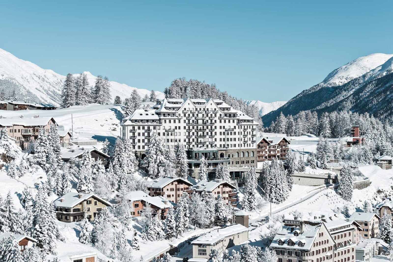 Carlton Hotel St. Moritz in winter: a white turreted building above snowy chalets and fir trees, with the Engadin mountains beneath a blue sky behind