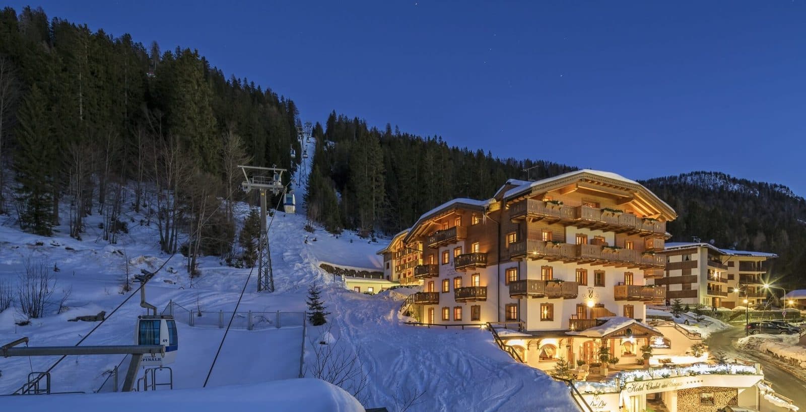 An illuminated wooden chalet beside a snowy slope with a lift and conifer forest