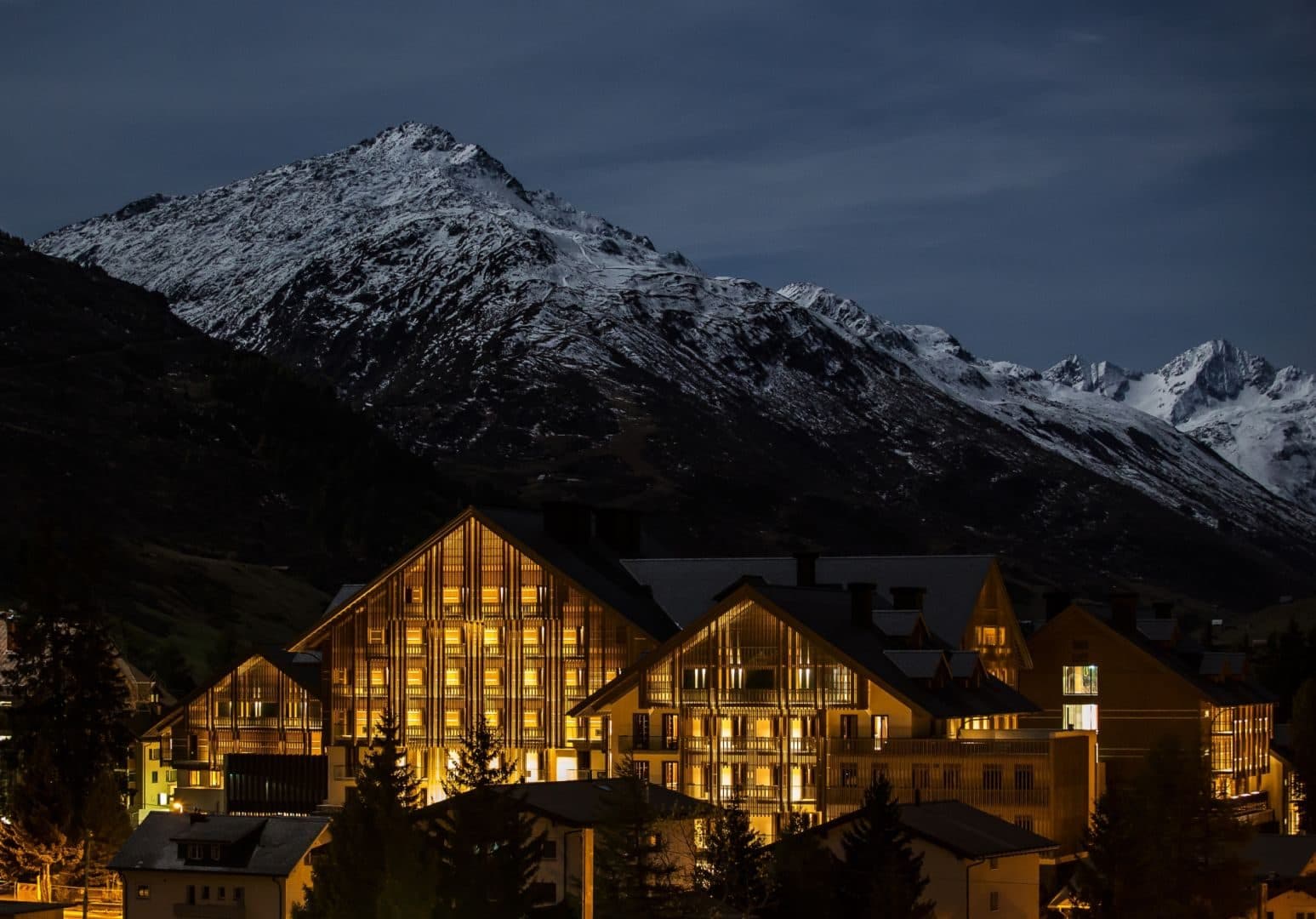 Illuminated buildings of The Chedi Andermatt in the evening beneath a snowy peak
