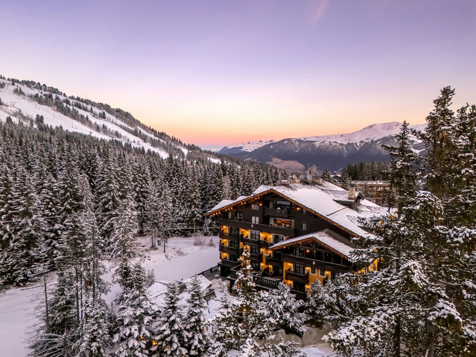 Aerial view of Cheval Blanc Courchevel at sunset: a dark wooden building with glowing windows among snowy fir trees, with a piste and mountains beneath a pink sky behind
