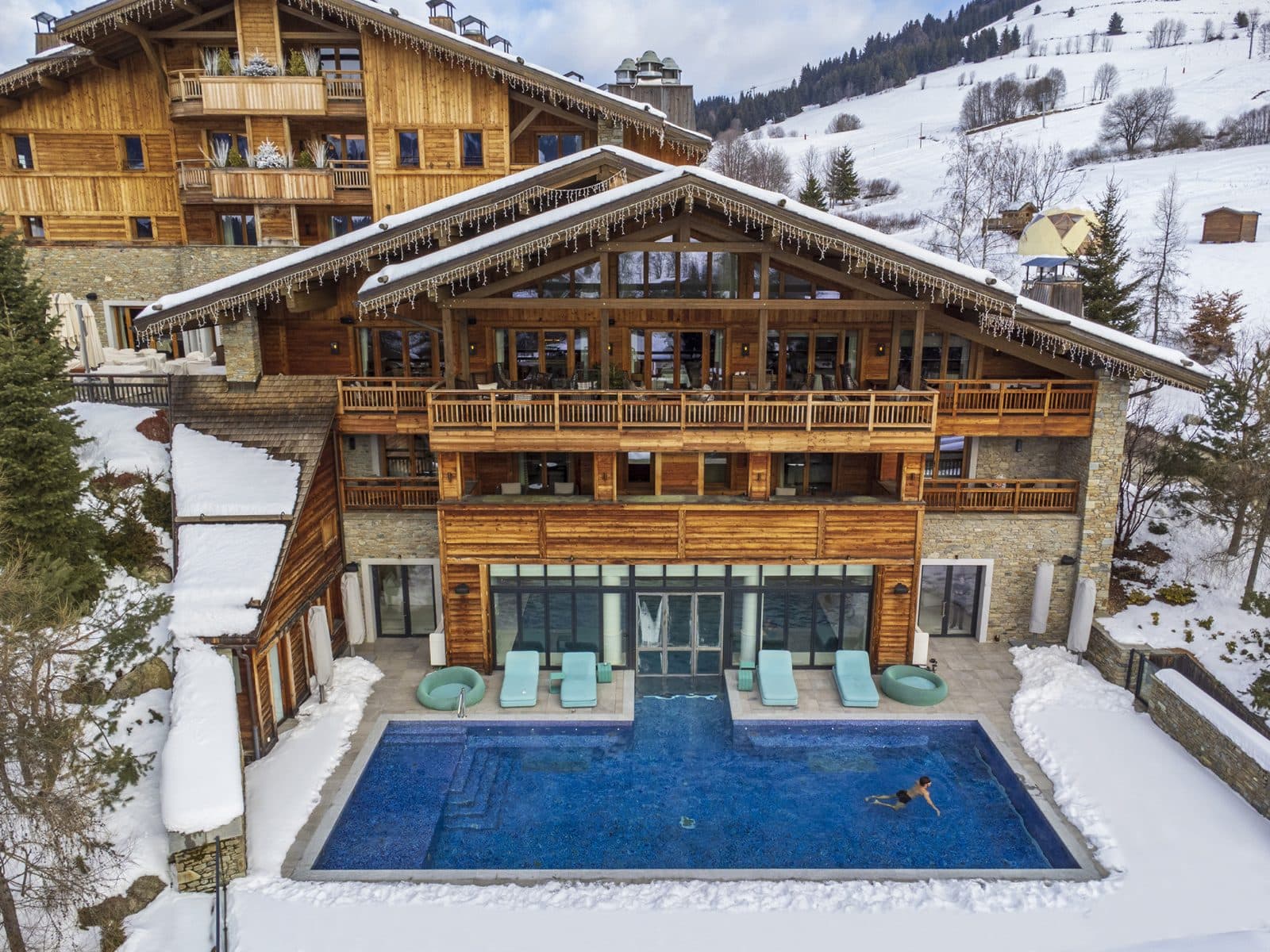 Aerial view of the wooden Four Seasons Megève chalets in snow: an outdoor pool with turquoise loungers before the spa’s glass façade