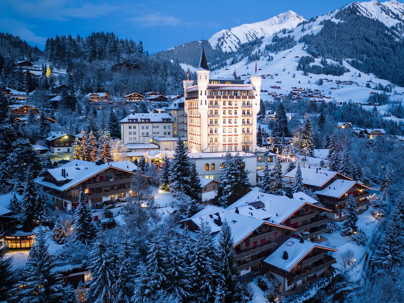 Gstaad Palace on a winter evening: a white castle with towers above Gstaad’s snowy chalets, with slopes and peaks behind