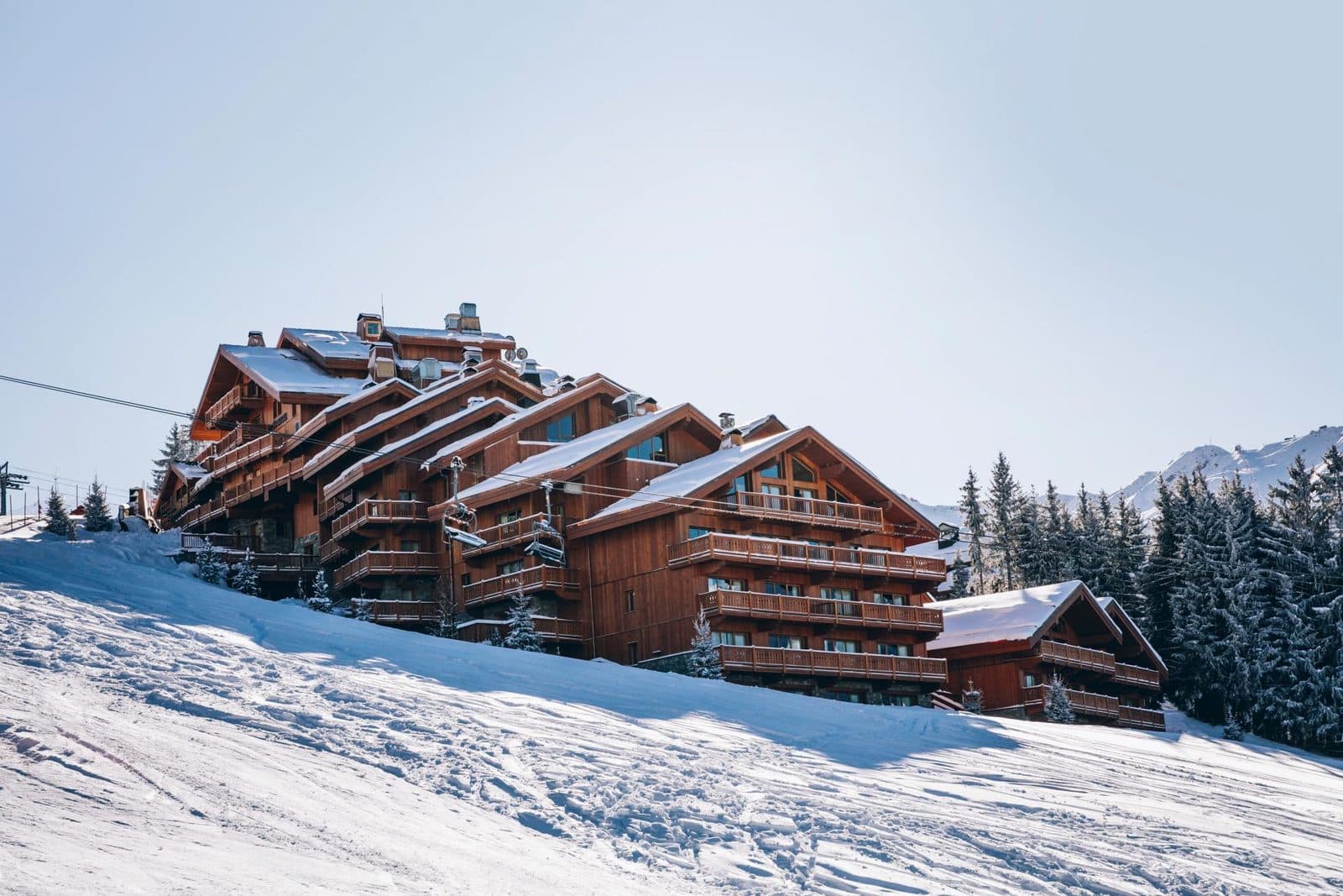 Le Coucou’s wooden building rises in tiers up the slope above a snowy piste, with a chairlift cable on the left and fir forest on the right