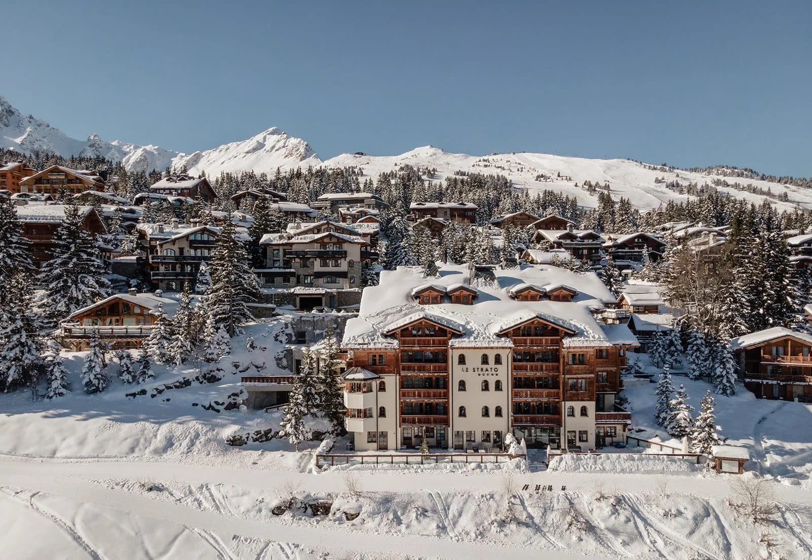Le Strato in snow: a pale façade with wooden balconies among Courchevel 1850 chalets, with snowy mountains behind