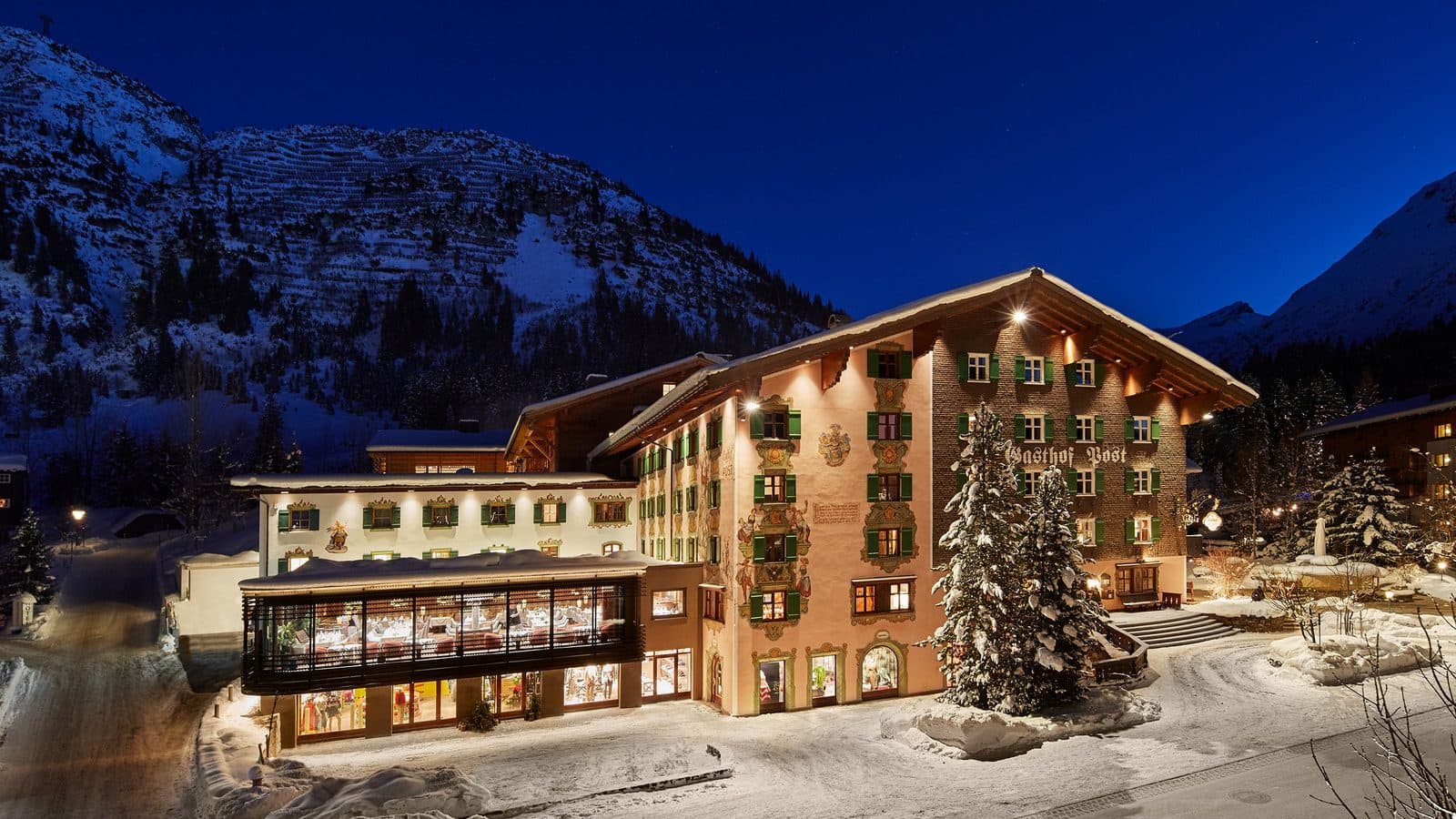 Hotel Post Lech in the evening: an illuminated façade with green shutters, snowy fir trees and mountains