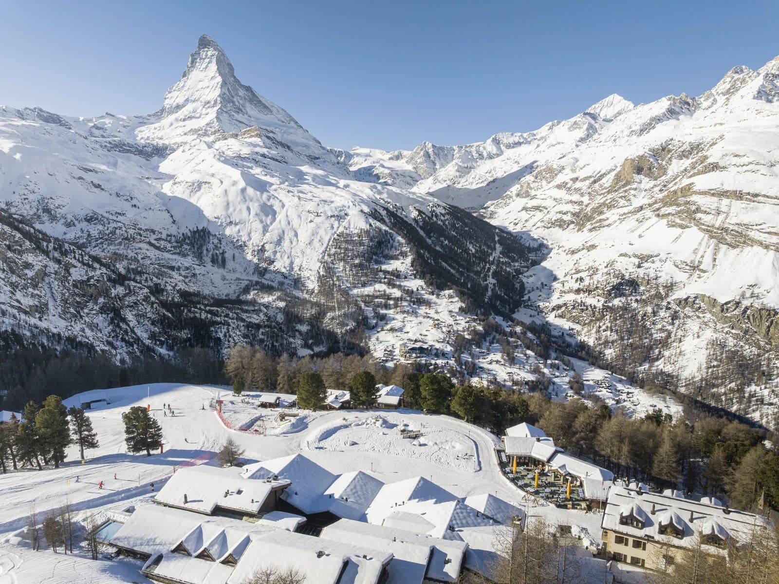 Aerial view of Riffelalp Resort in winter: snowy hotel roofs on a plateau among larches, with the Matterhorn and Zermatt valley behind