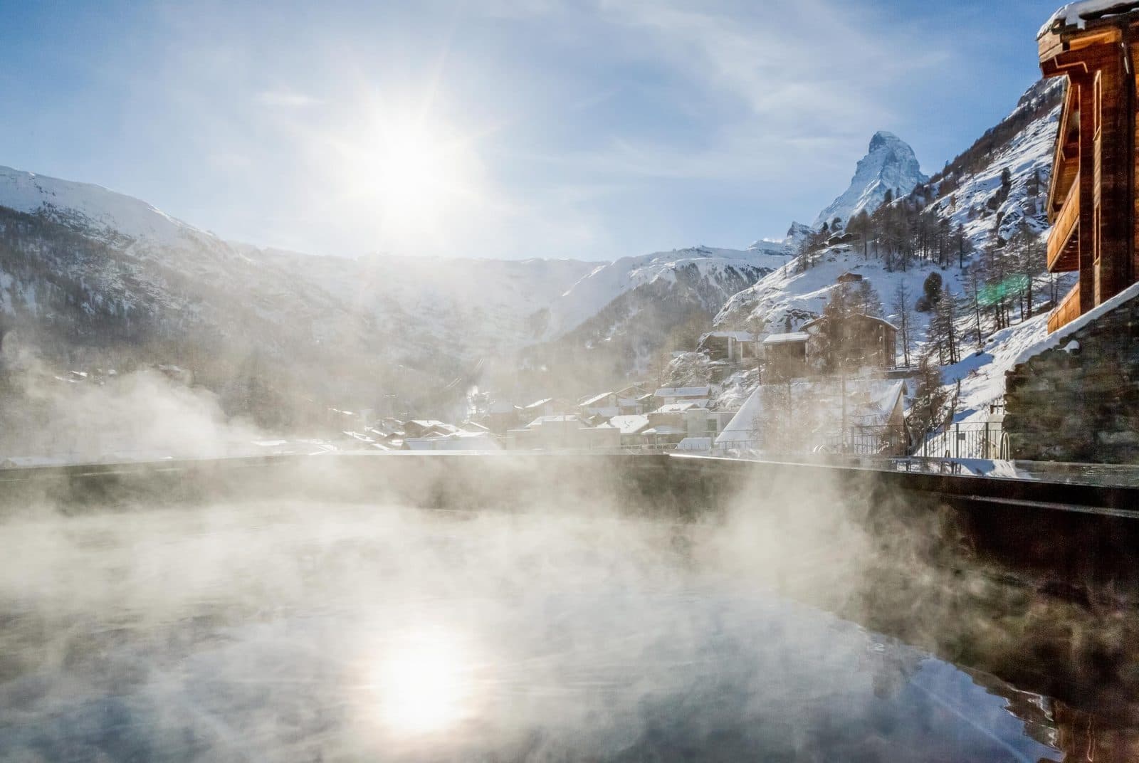 The Omnia’s outdoor pool in winter: steam above the water, the snowy Zermatt valley and Matterhorn beyond, and the hotel’s wooden façade on the right
