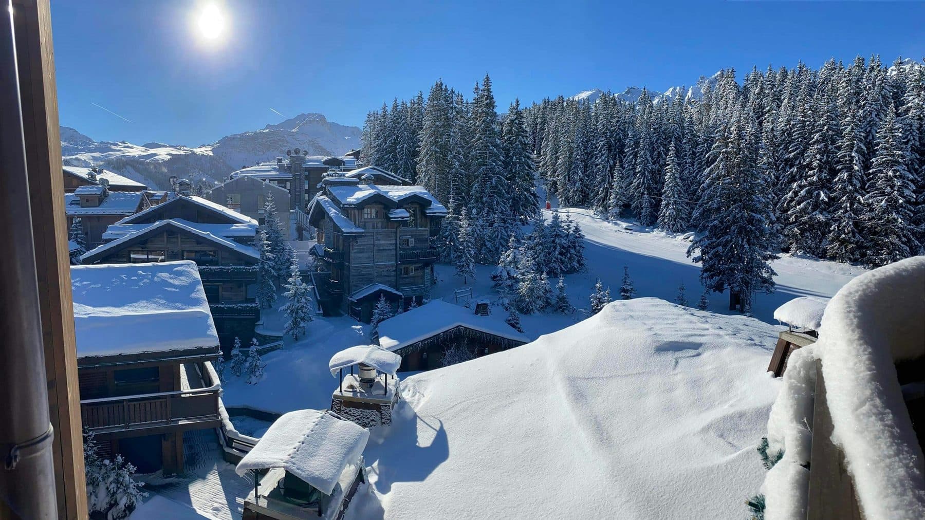 View from a balcony in Courchevel: chalets beneath thick snow among fir trees, with mountains in the distance and sunshine in a blue sky