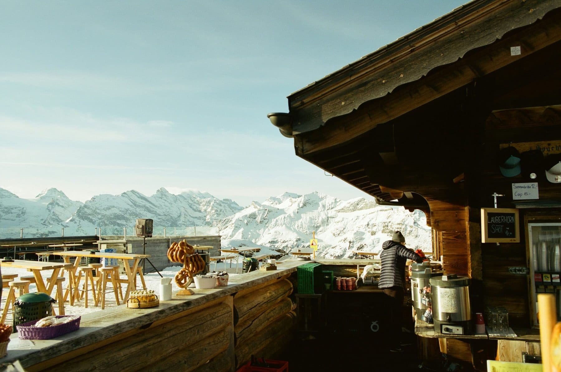 A sunny mountain bar terrace on Allmendhubel above Mürren: a wooden counter with pretzels, tables in the snow and a panorama of the snowy Alps