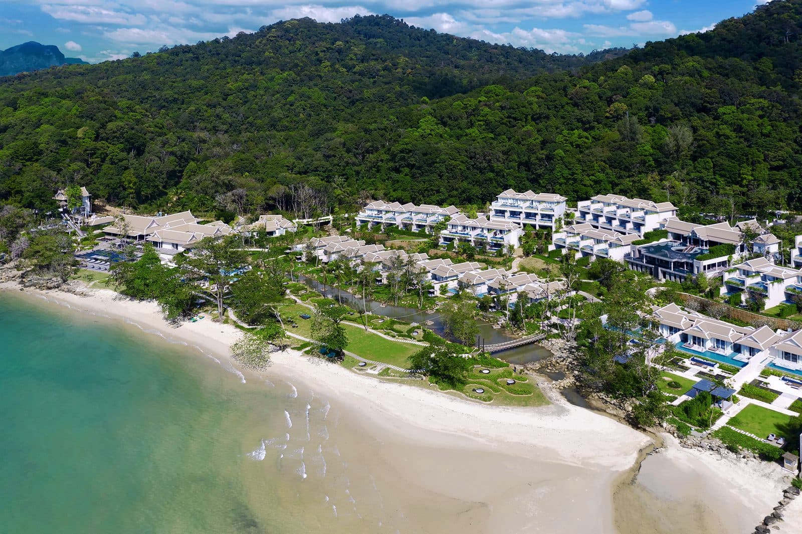 Aerial view of Banyan Tree Krabi: white buildings and villas among gardens by Tubkaek Beach, wooded mountains above the resort and turquoise water