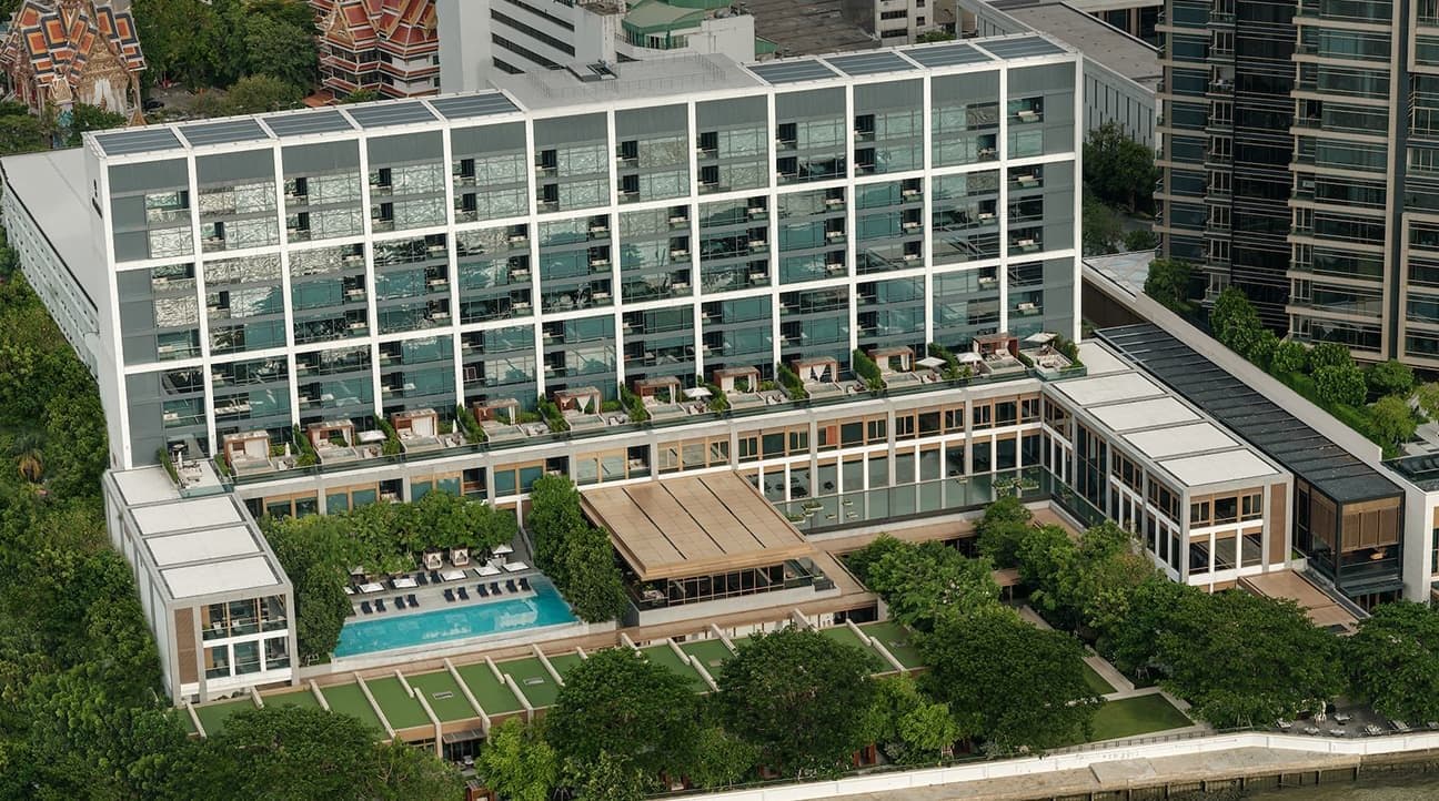 Aerial view of Capella Bangkok: a low building with a glass façade and terraces, pool and lawns in a Chao Phraya riverfront garden, with Thai temple roofs beyond