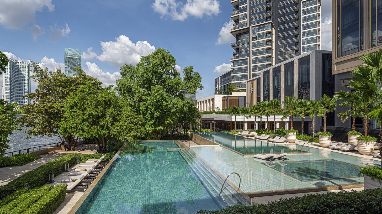 Two infinity pools cascade towards the Chao Phraya: loungers, potted palms, old riverside tree canopies and hotel towers