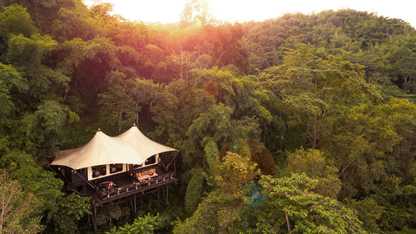 A tent on a wooden platform amid dense hillside jungle, with the sun setting above the hill