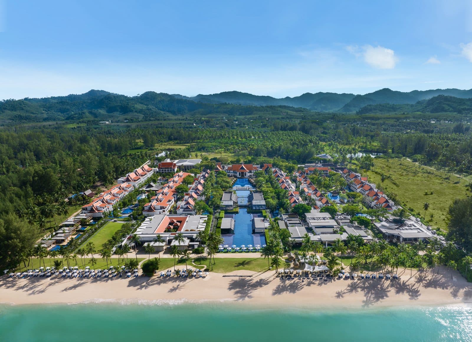 Aerial view of the resort by Khuk Khak Beach: red-roofed buildings, a lagoon pool across the grounds, beach loungers and jungle mountains on the horizon