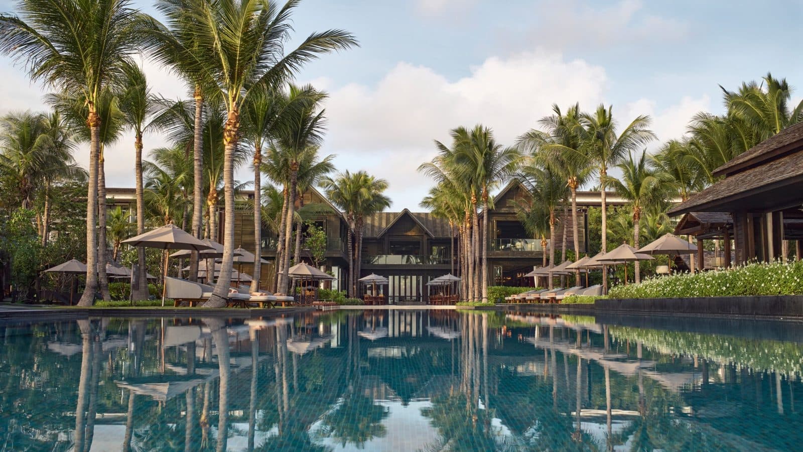Kimpton Kitalay Samui's main pool: coconut palms, umbrellas and dark pitched-roof buildings reflected in still water