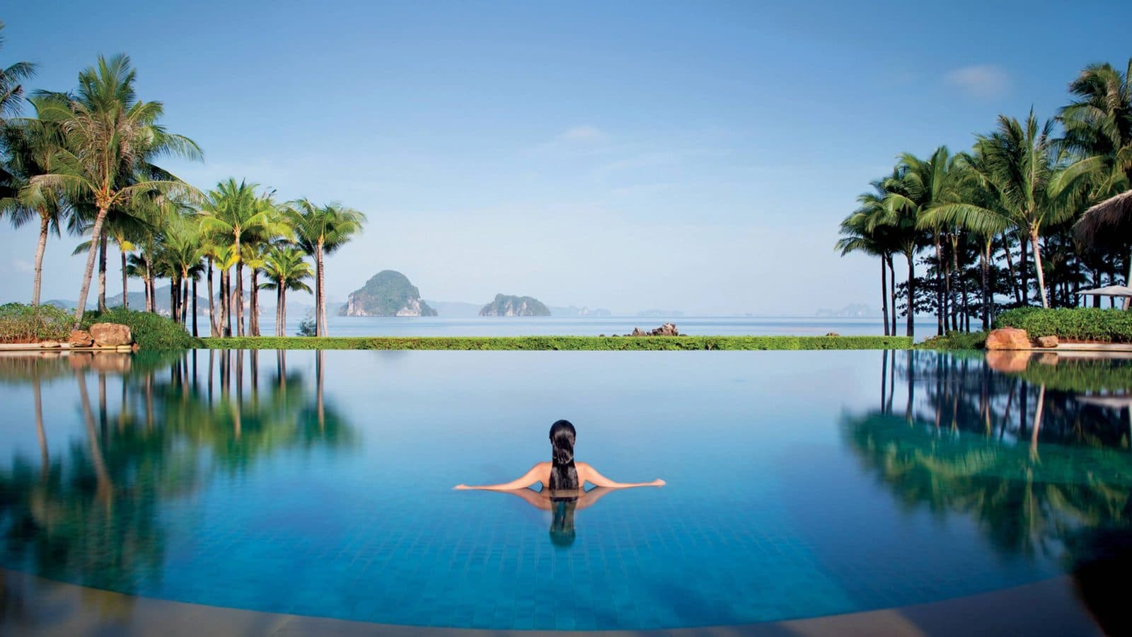 An infinity pool between palms faces the Andaman Sea and limestone islands; a woman at the pool edge has her back to the camera
