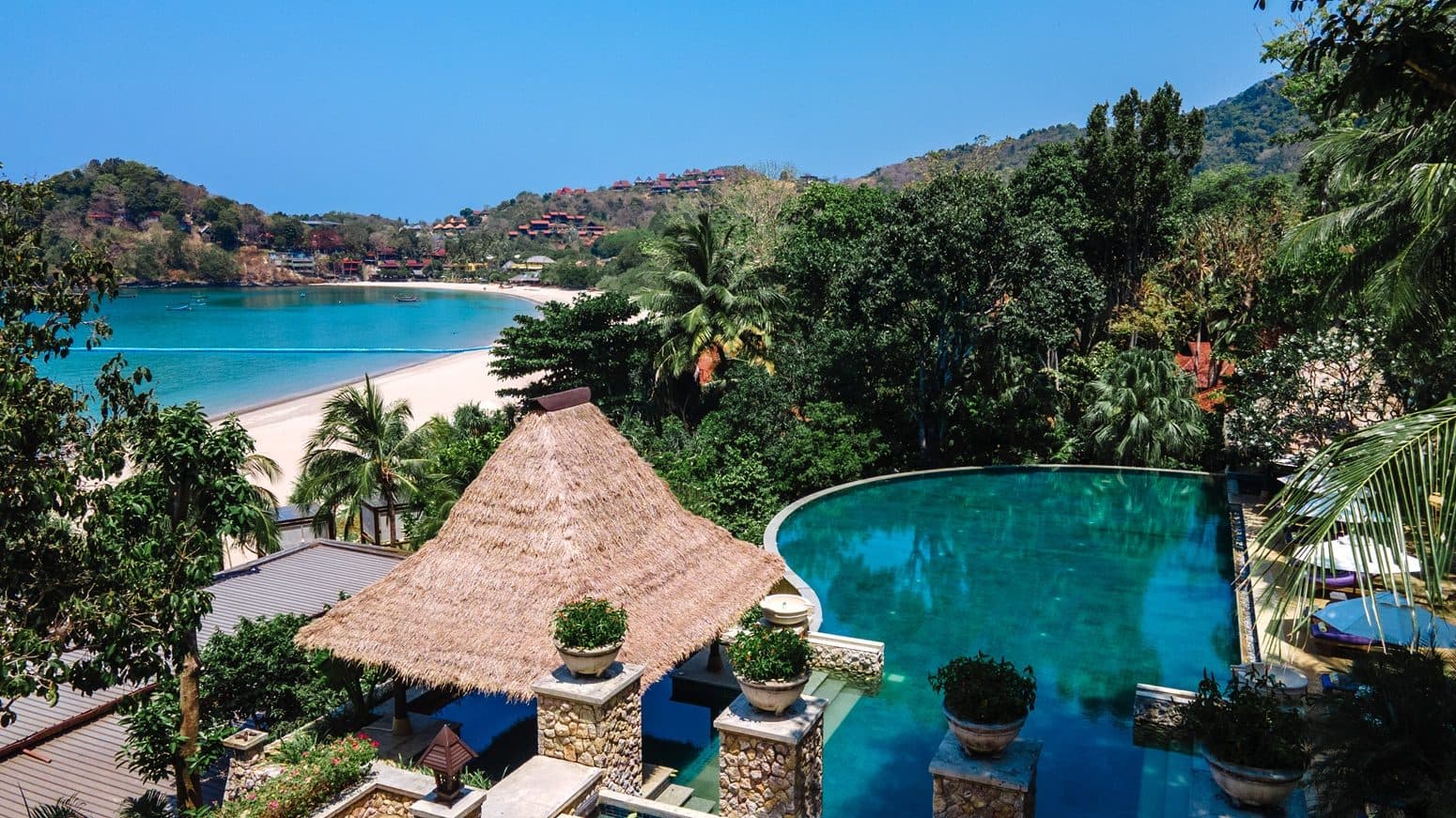 Aerial view of Pimalai's infinity pool and the thatched gazebo above it. Beyond the palms, a bay with a white beach and houses on green hills