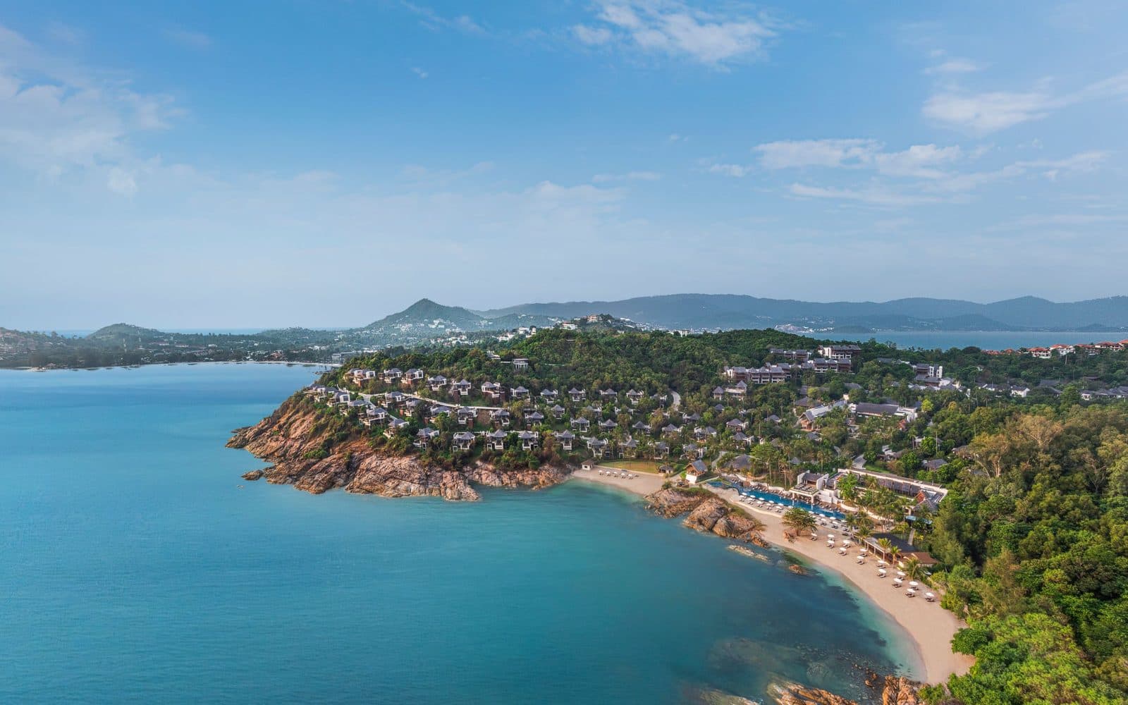 Aerial view of The Ritz-Carlton, Koh Samui: villas on a rocky headland, a beach with a bay-side pool and Samui's mountains in the distance