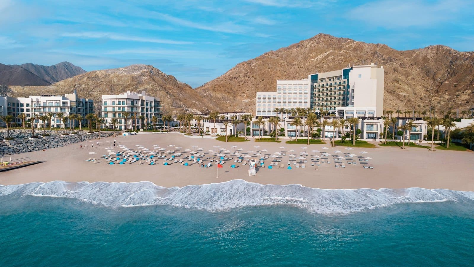 Address Beach Resort Fujairah from the sea: a broad beach with umbrellas and loungers, white hotel buildings among palms and the Hajar Mountains behind