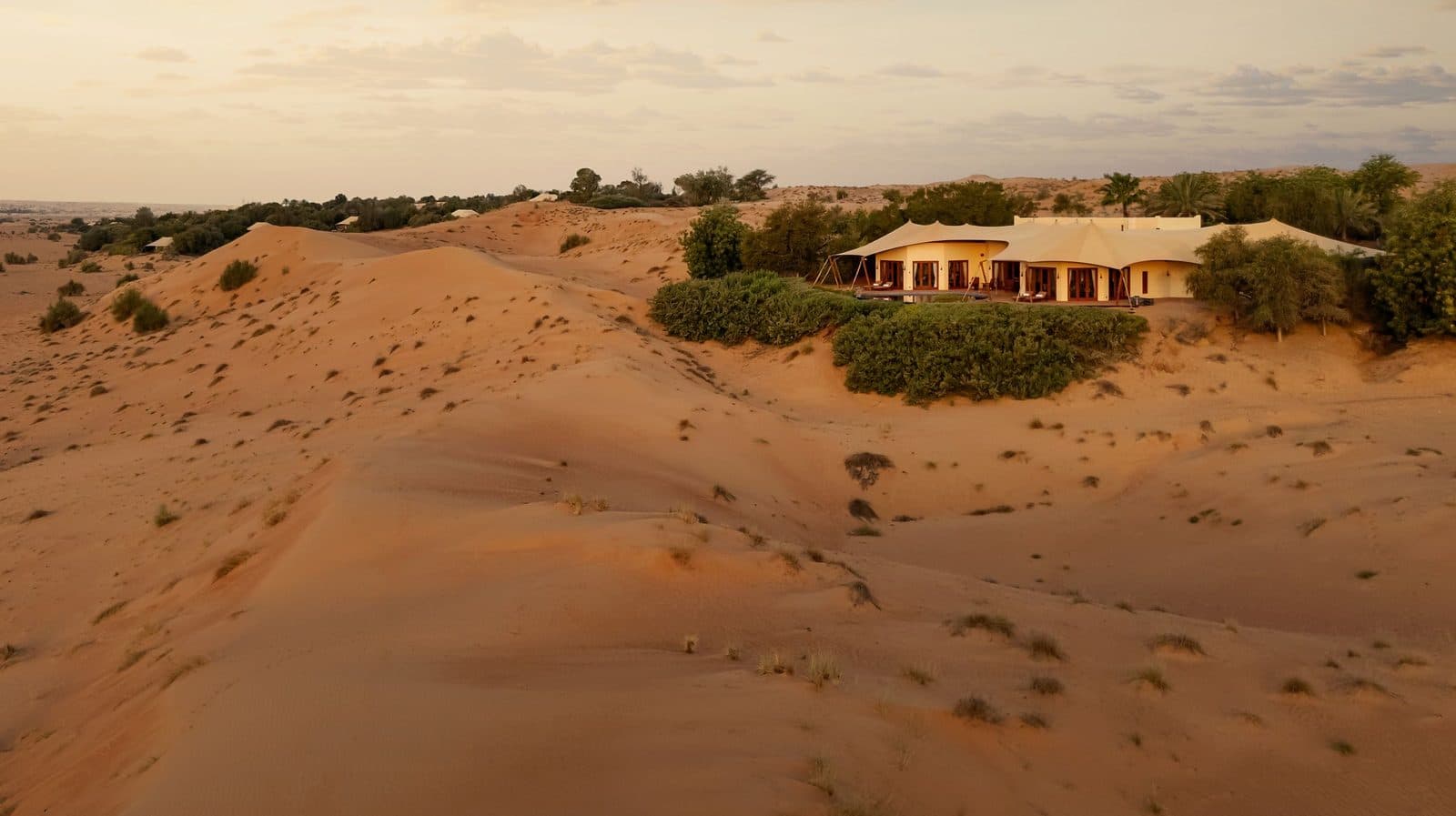 An Al Maha villa among dunes at sunset: tented roofs amid greenery, rippled sand and scrub stretching to the horizon