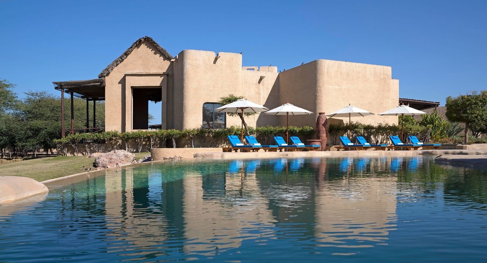 Al Sahel’s free-form main pool reflecting a sand-coloured thatched building, with blue loungers and white umbrellas