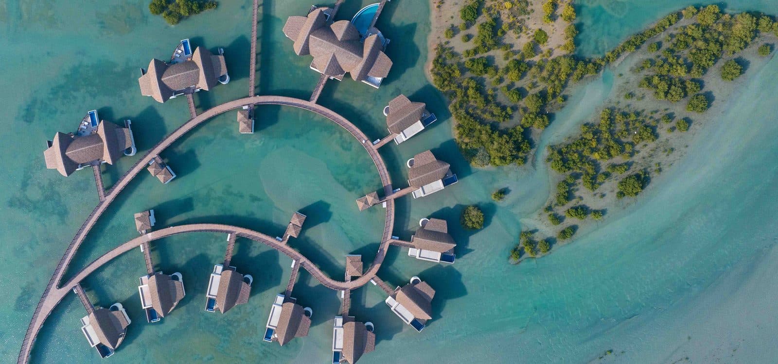 Aerial view of Anantara Mina Ras Al Khaimah’s overwater villas: curved wooden walkways, houses with pools and mangroves in a turquoise lagoon