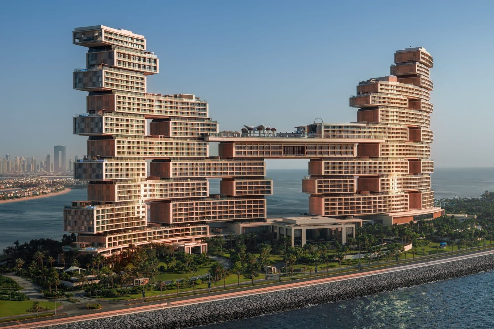 Atlantis The Royal from the air: two stepped block towers linked by a bridge, a palm garden and waterfront by the breakwater; Palm fronds and Dubai Marina skyscrapers in the distance