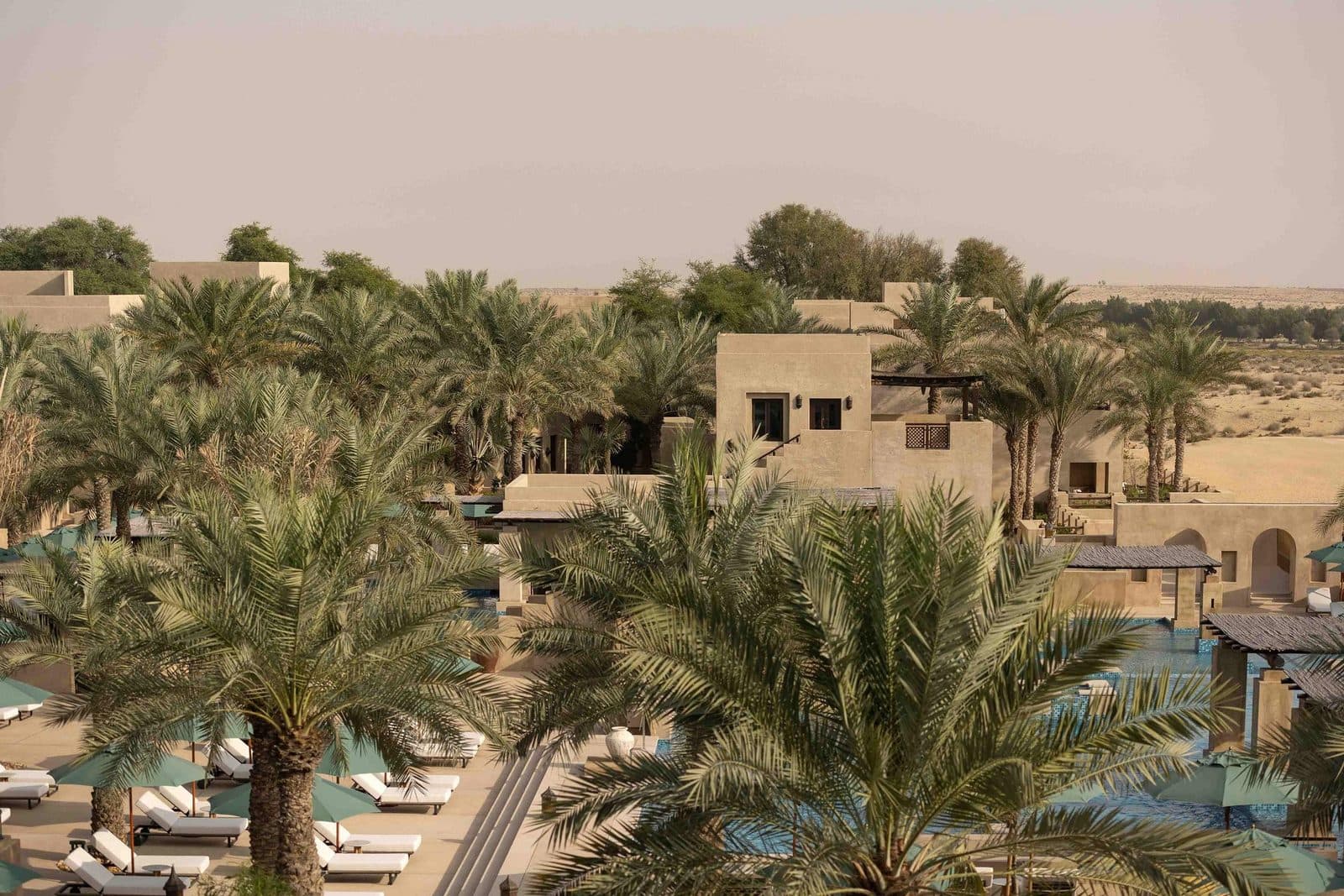 Bab Al Shams from above: sand-coloured buildings among date palms, a pool with sun loungers and desert beyond the resort
