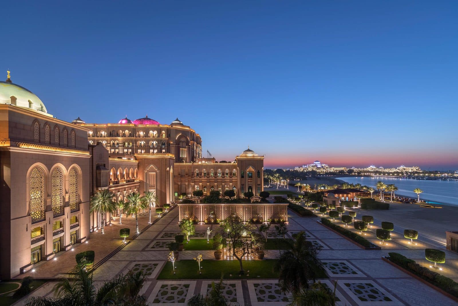 Emirates Palace at dusk: illuminated arches and domes, a garden with patterned paths and a beach beside the Gulf