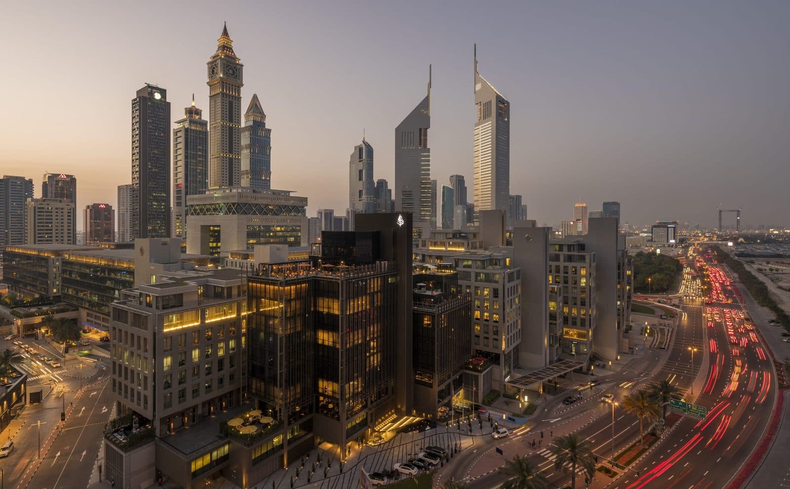 Aerial evening view of DIFC: a dark-glazed hotel amid Gate Village, with Emirates Towers behind and traffic on the right