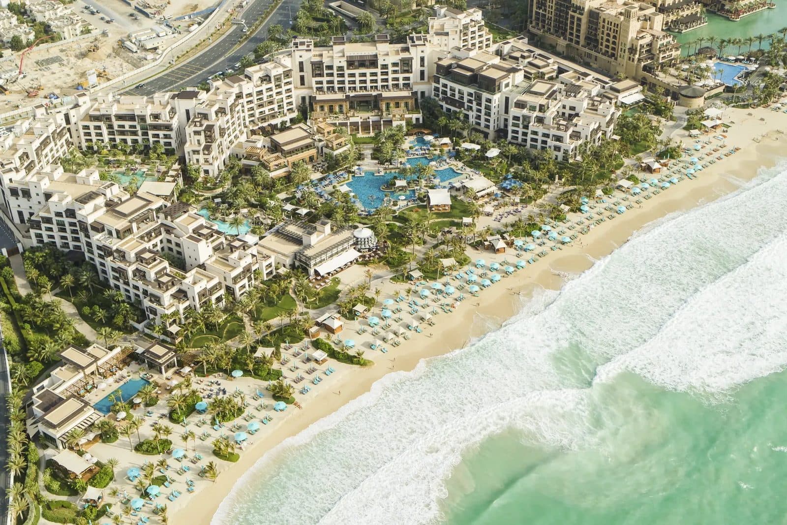 Jumeirah Al Naseem from above: pale buildings among palms and pools, a beach with light-blue umbrellas and white surf
