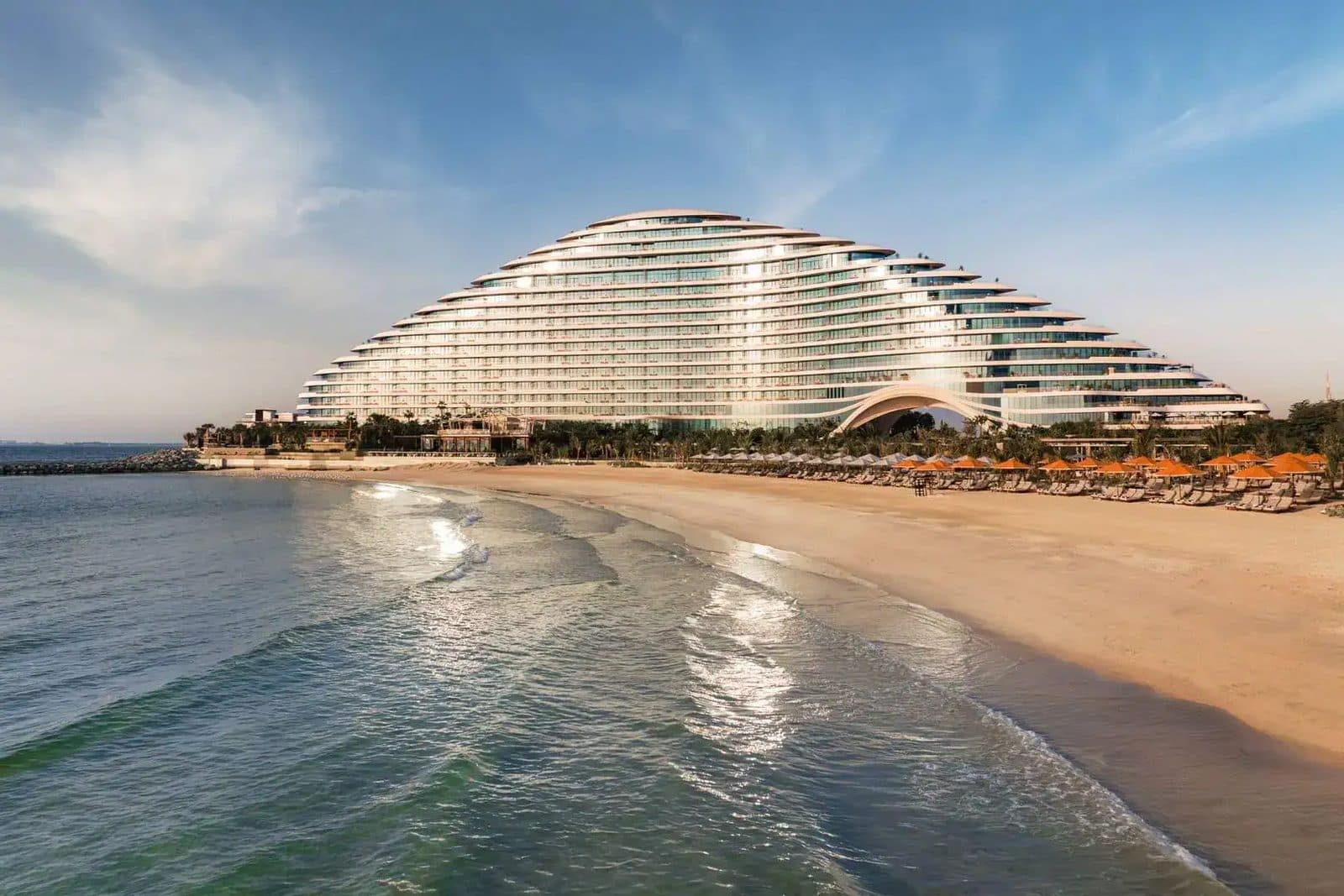 Jumeirah Marsa Al Arab from the sea: a wave-shaped white building behind a broad sandy beach with orange umbrellas
