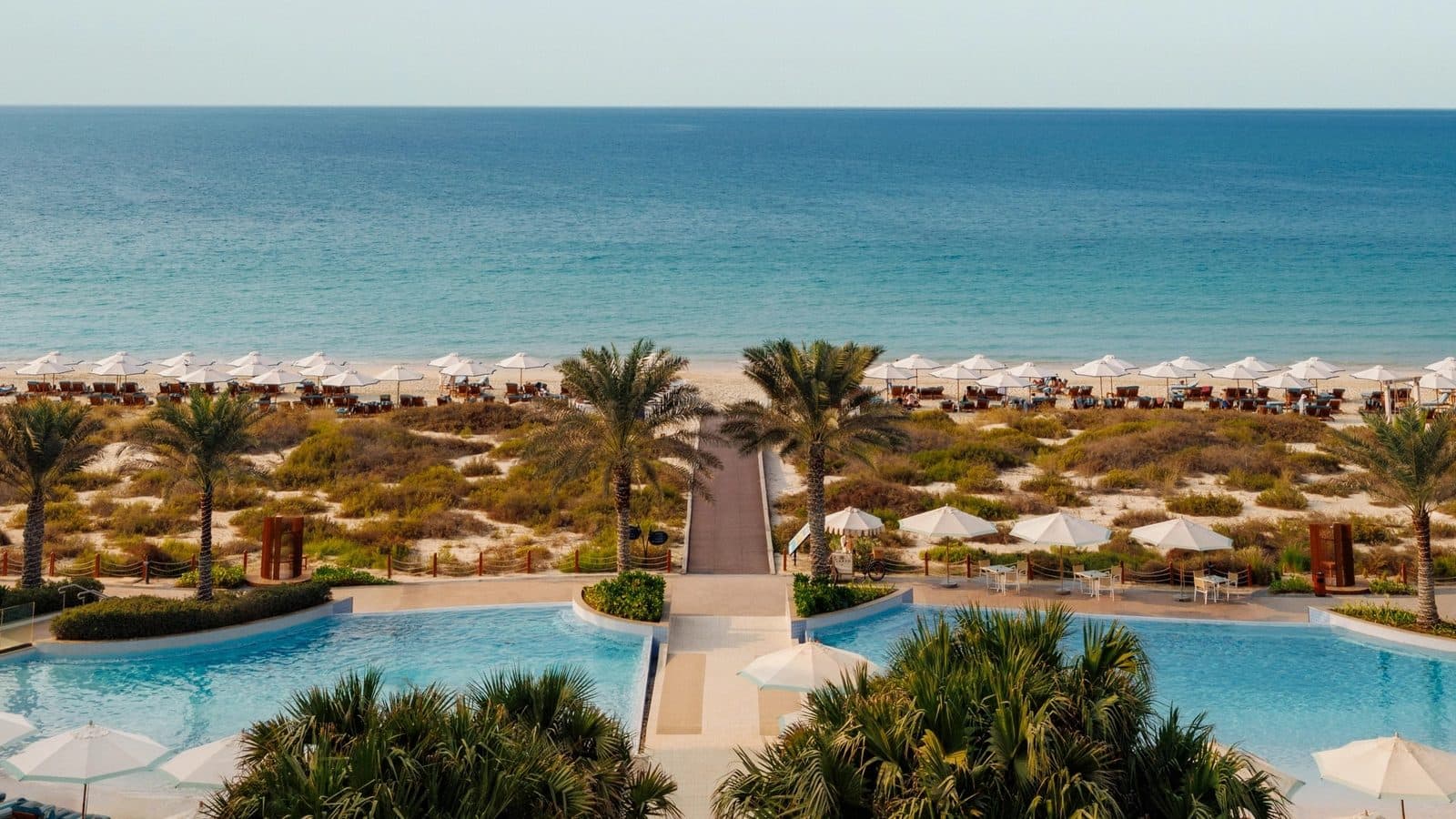 View from Jumeirah Saadiyat Island’s terrace: two pools among palms, a wooden path through dunes to a beach with white umbrellas and the Persian Gulf