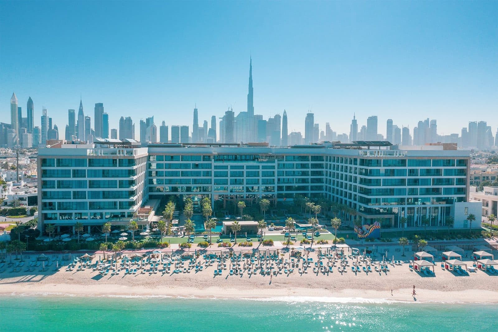 Mandarin Oriental Jumeira viewed from the sea: a beach with umbrellas and cabanas, garden and hotel building, with skyscrapers and the Burj Khalifa beyond