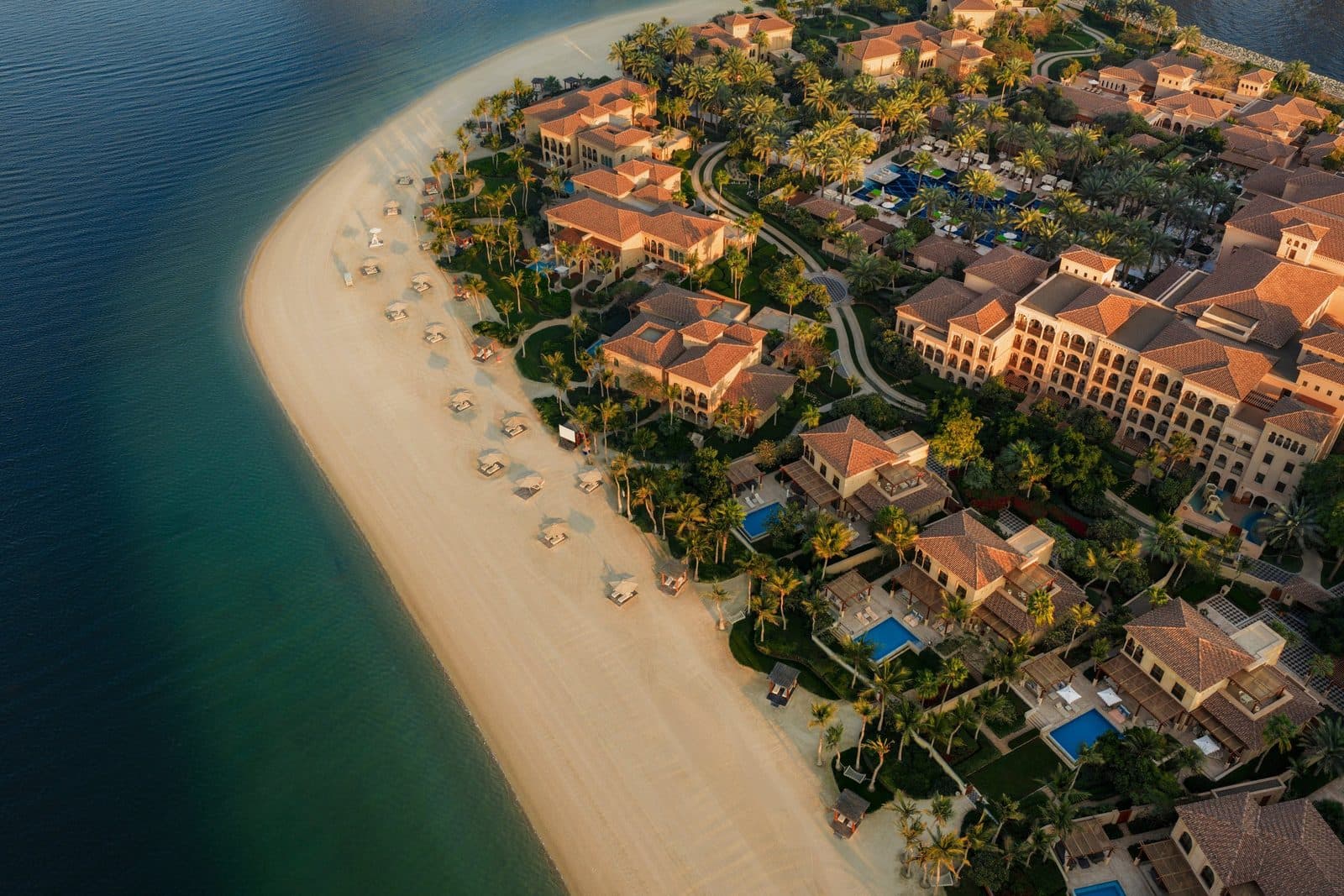 Aerial view of One&Only The Palm in evening light: a curved beach with umbrellas, private-pool villas in a palm garden and Andalusian buildings with tiled roofs