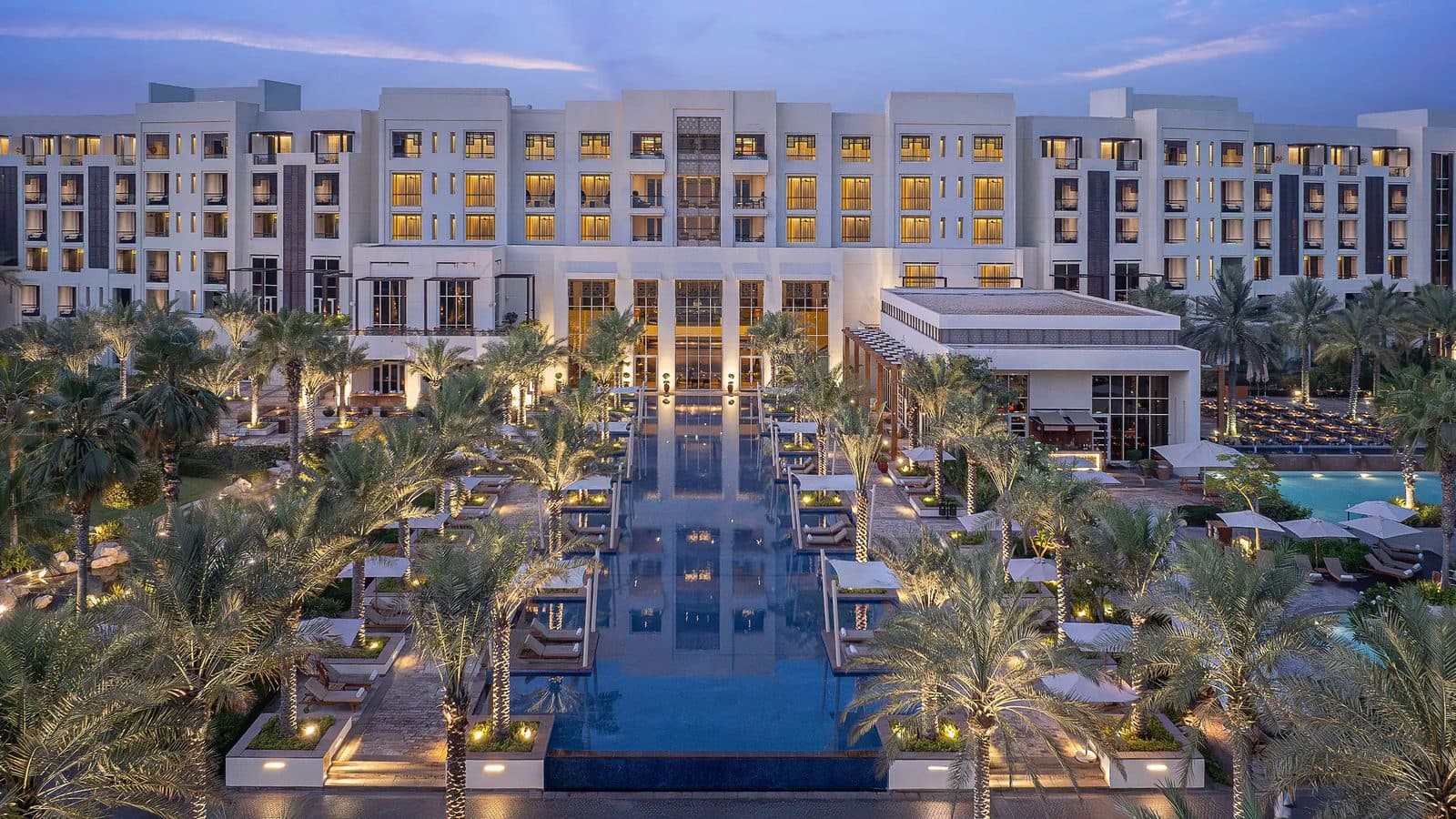 Park Hyatt Abu Dhabi at dusk: a long pool with sun loungers between palms leading towards the pale hotel building