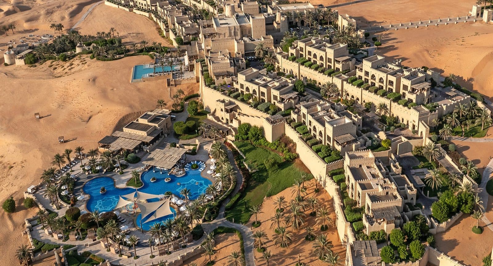 Aerial view of Qasr Al Sarab: sand-coloured buildings with towers and crenellated walls, palm gardens and a pool with tents among dunes