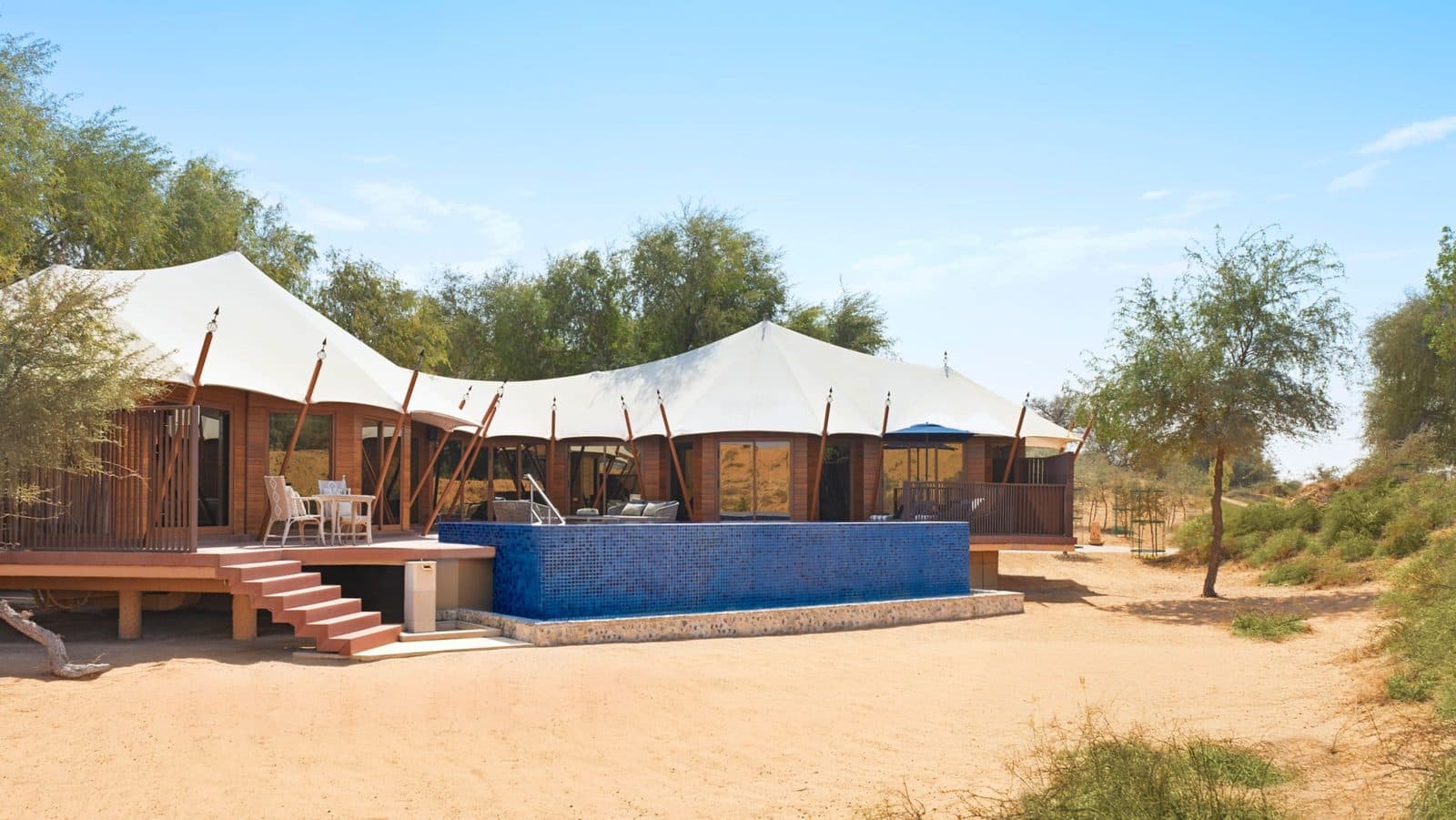 An Al Sahari tented villa: white tented roofs, a stepped wooden terrace and a blue-mosaic pool among trees on sand