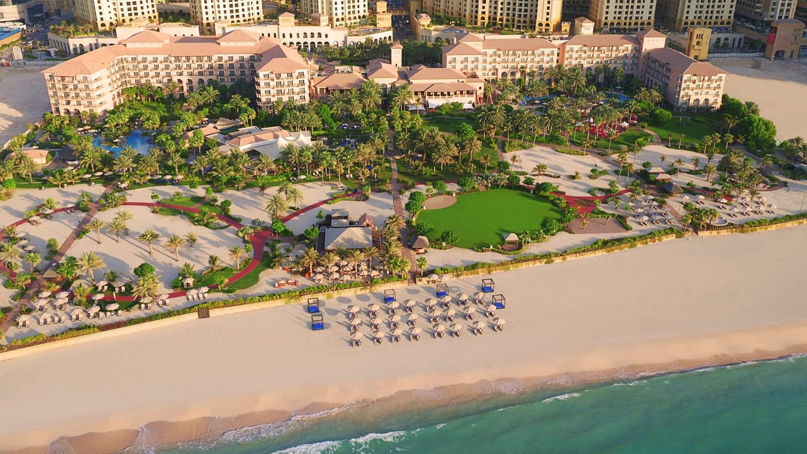 The Ritz-Carlton, Dubai from above: terracotta-roofed buildings among palms and lawns, a broad beach with rows of umbrellas and turquoise sea