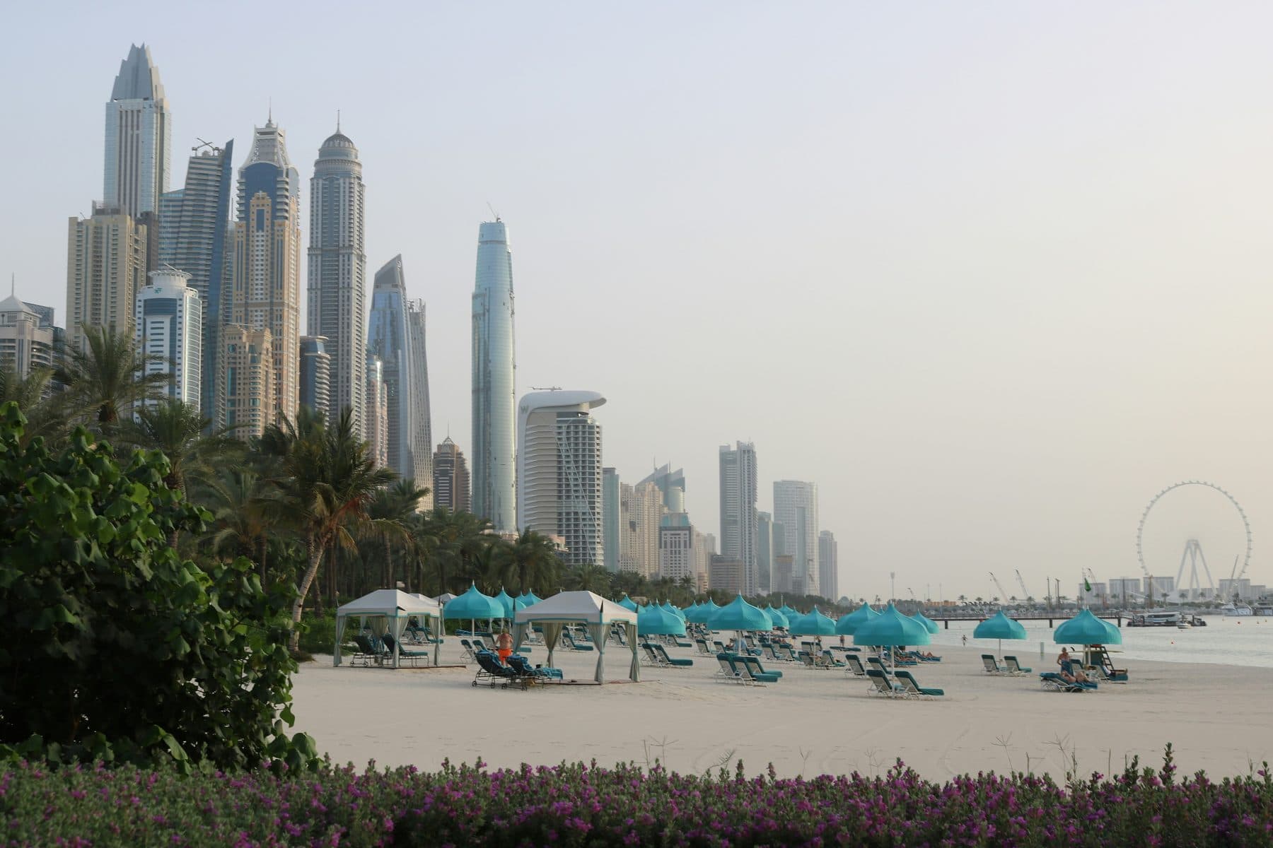 A beach with turquoise umbrellas, sun loungers and white tents; Dubai Marina towers behind the palms, with a Ferris wheel in the distance on the right