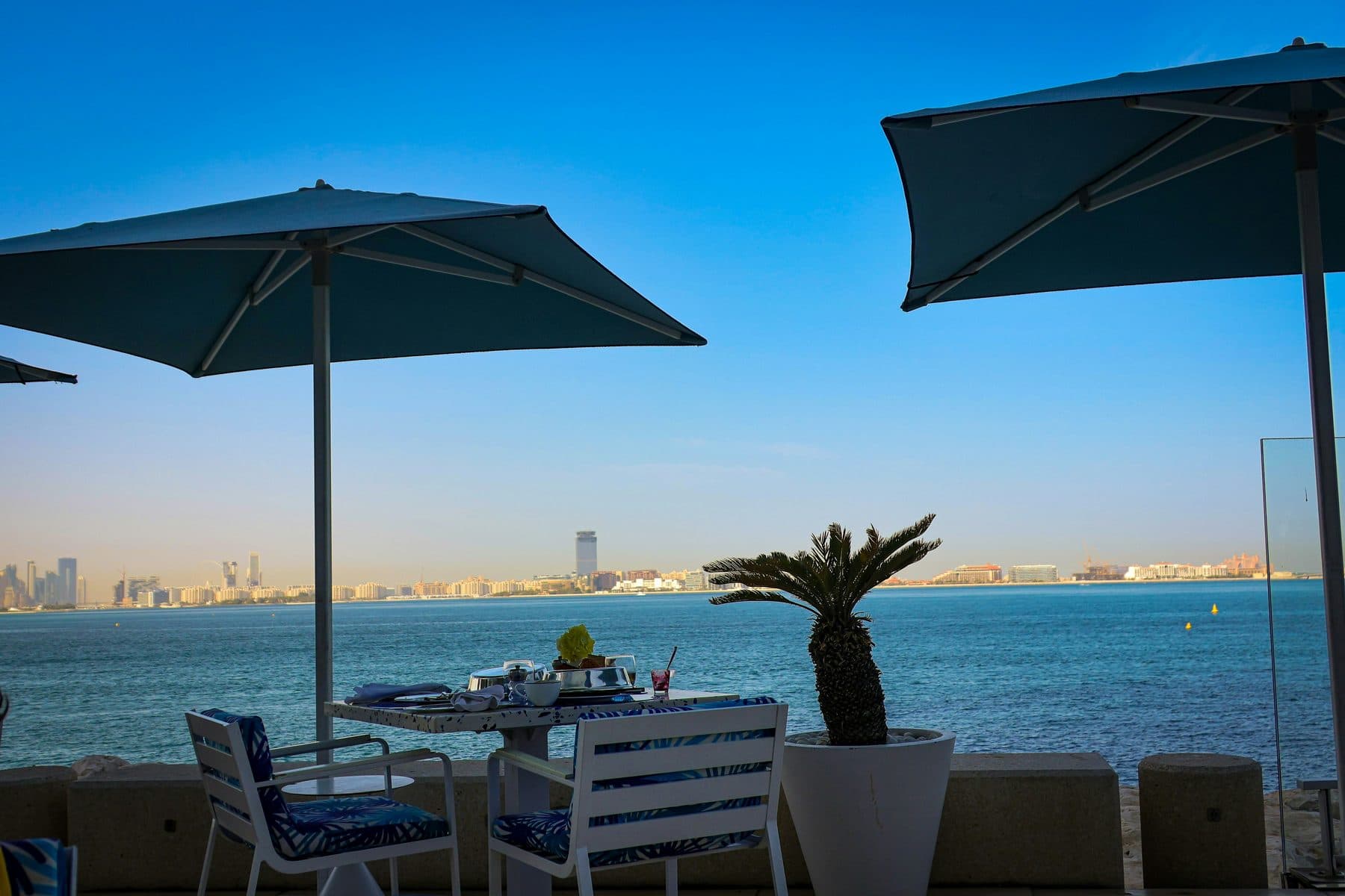 A set table with white chairs beneath dark umbrellas on a terrace beside the water; Dubai’s skyscraper-lined shore across the Gulf
