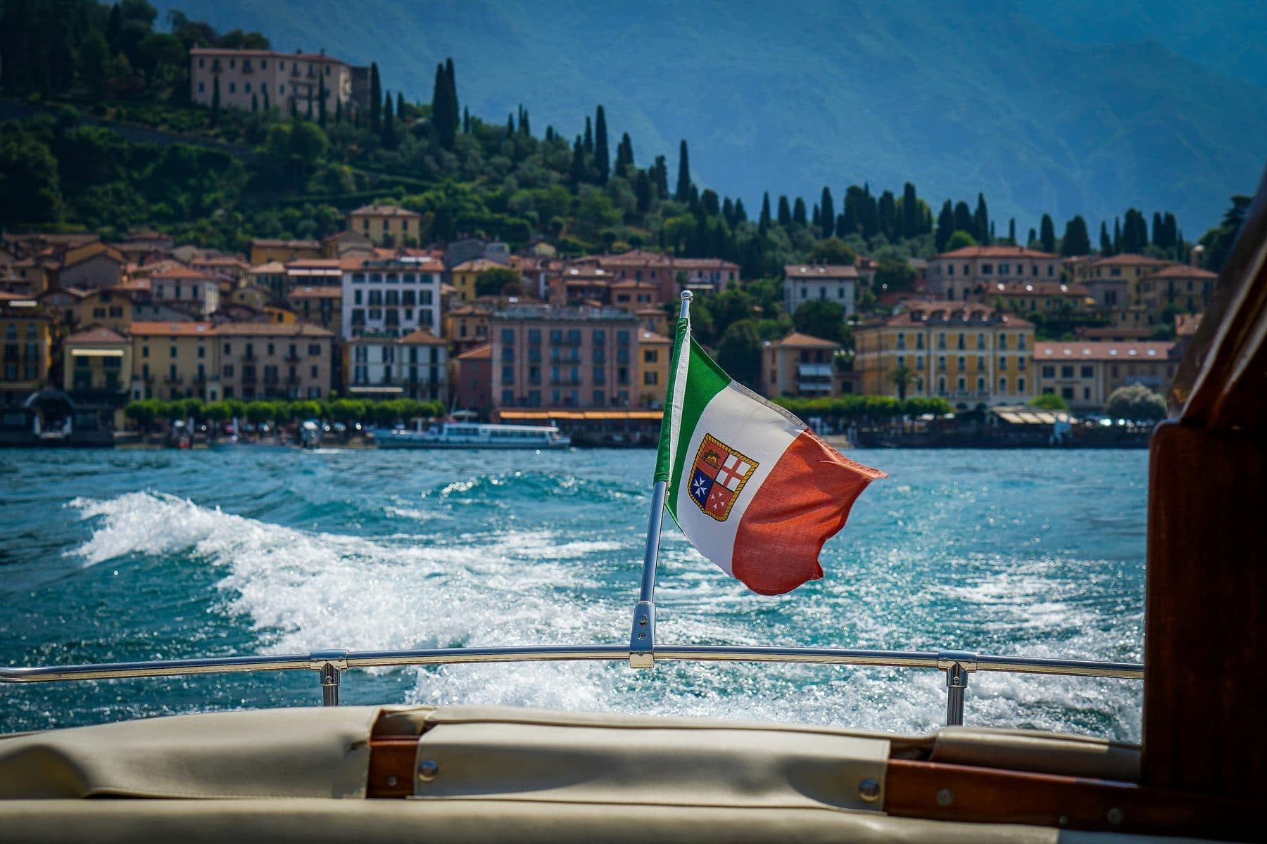 View from the stern of a wooden boat on Lake Como: an Italian flag, white wake and a town waterfront with houses and cypresses