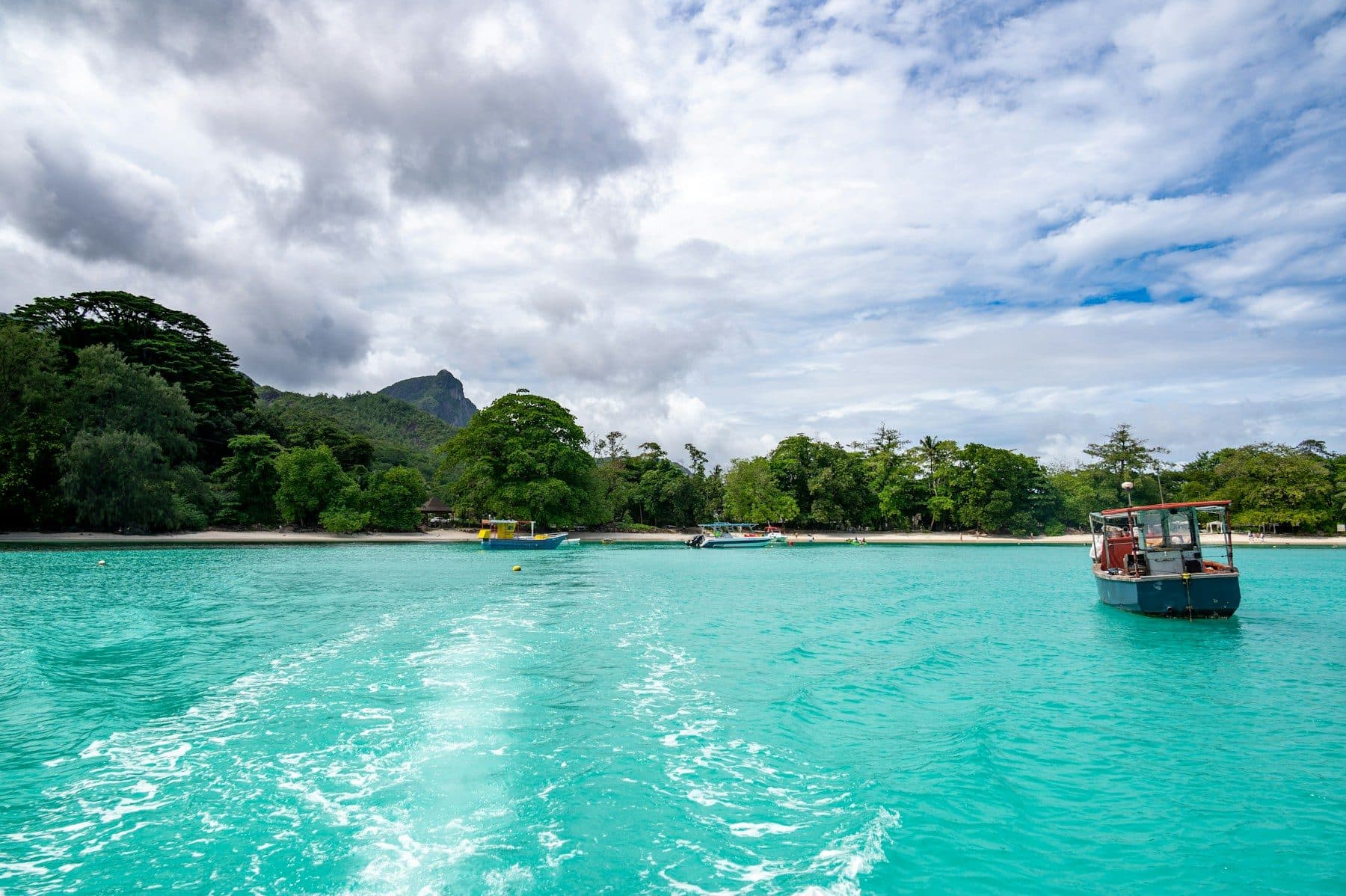 A speedboat's foamy wake on turquoise water off Mahé: a beach and forest with a granite peak ahead, and a fishing boat anchored on the right