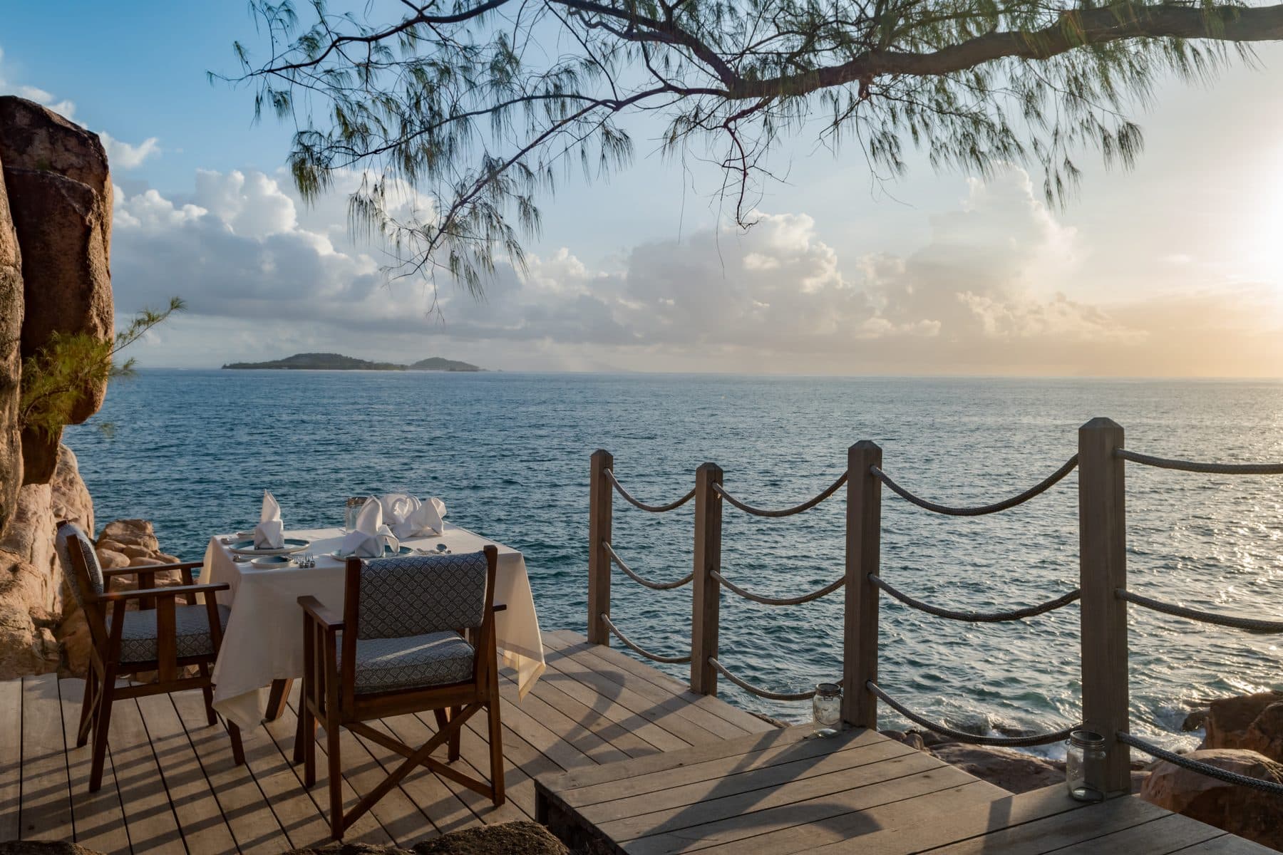 A white-clothed table for two on a wooden terrace above the sea on Praslin: rope railings, a low sun and islands on the horizon