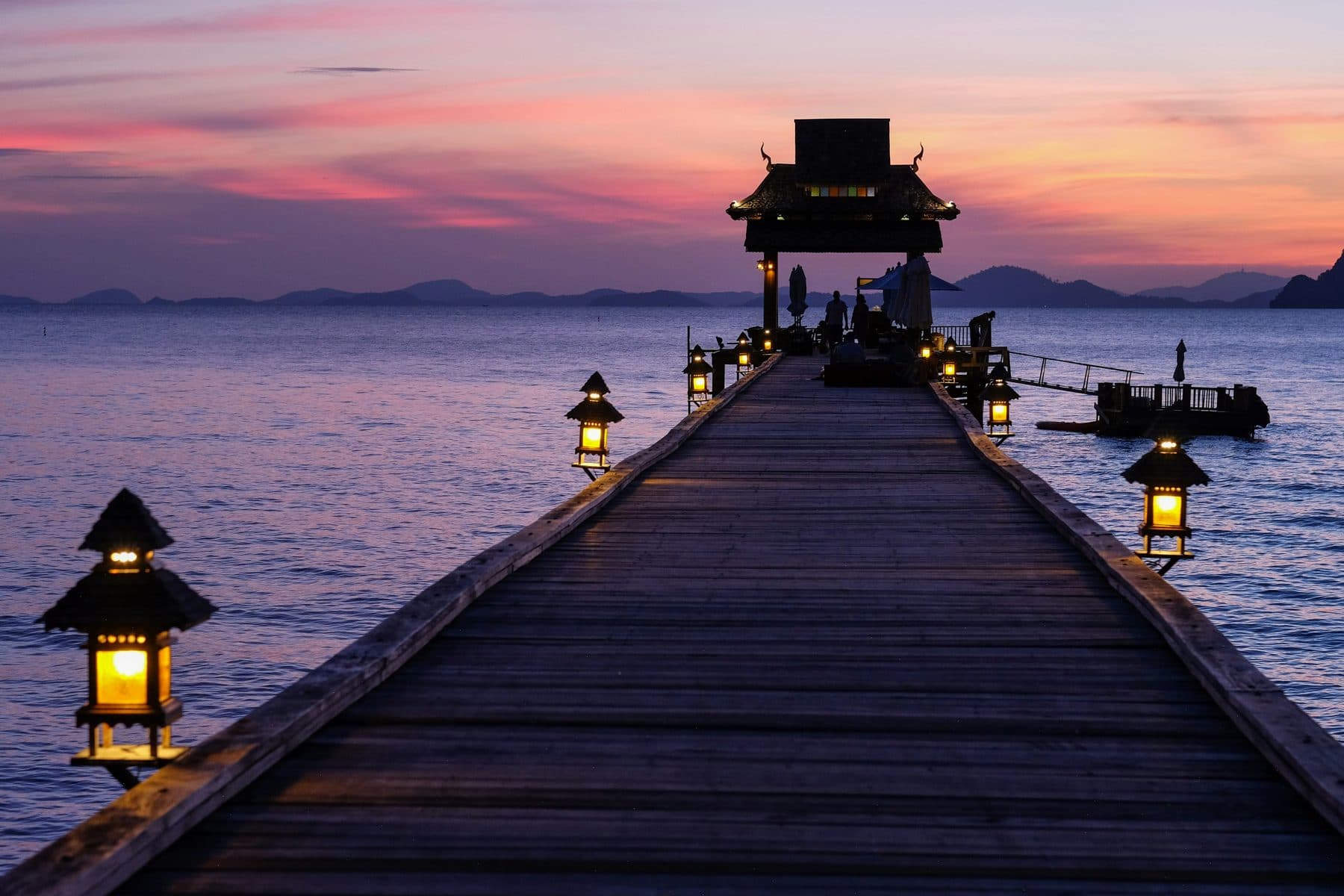 A wooden pier with lit lanterns leads to a Thai-roofed gazebo above the sea by Yao Yai island; silhouettes beside the gazebo, a pink post-sunset sky and distant islands